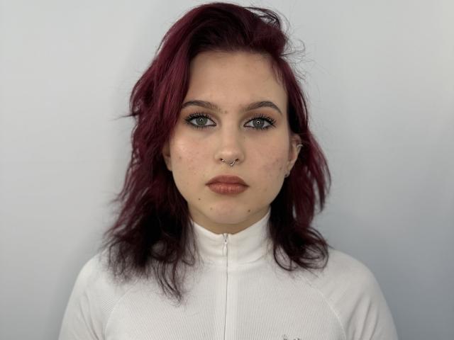 SheritaPinales tiene cabello rojo, ojos verdes, un piercing en la nariz y una camisa blanca con cremallera. Está mirando directamente a la cámara con una expresión seria. Sus senos no son visibles..