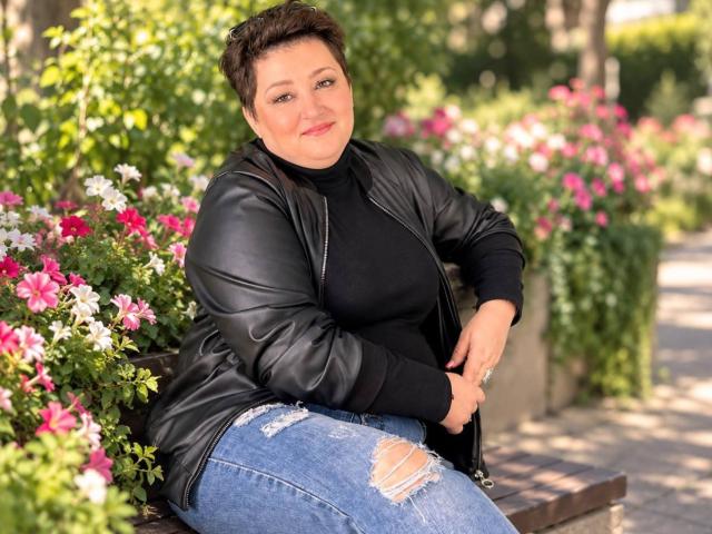 LouiseGrayy, con cabello castaño corto, un suéter negro de cuello alto, una chaqueta de cuero negro y jeans azules rotos, sentada en un banco, sonriendo, rodeada de flores rosas y blancas..