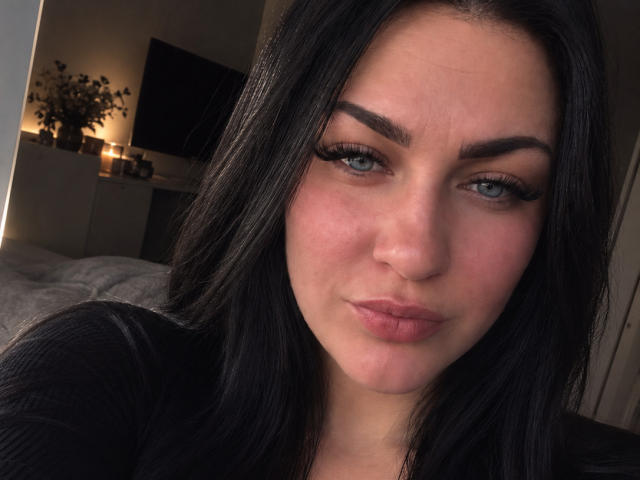 KarolineFran has puffy pink lips, thick black eyebrows, long black hair, and blue eyes with long eyelashes. She's wearing a black top, looking slightly annoyed.