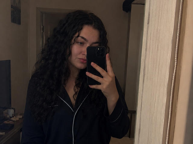 KarolineFran, com cabelo preto encaracolado, vestindo uma jaqueta preta com zíper, tirando uma selfie no espelho com um celular preto, com a mão cobrindo parte do rosto, em iluminação fraca..