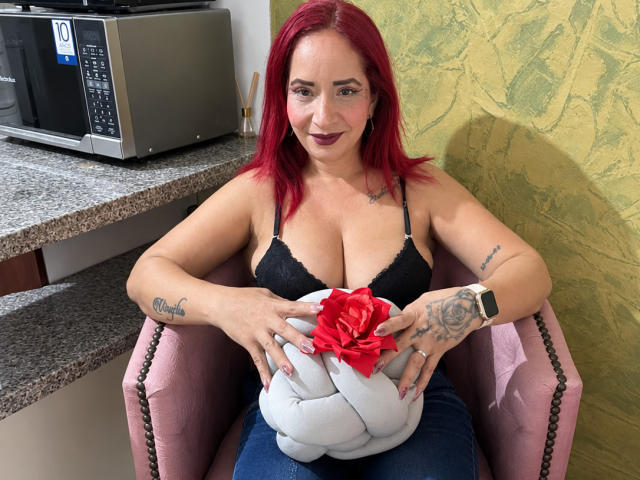 KateMonroe with red hair, black bra, tattoos, holding white gift with red bow, sitting in pink chair, wearing watch, looking at camera, in kitchen, microwave on counter.