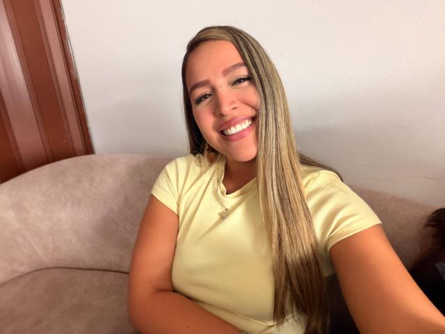 LiaDiane, con lunghi capelli biondi, una camicia gialla, che sorride mostrando i denti, seduta su un divano beige, con la pelle abbronzata, piccoli seni, dall'aspetto rilassato, un selfie casual, con una porta di legno sullo sfondo..