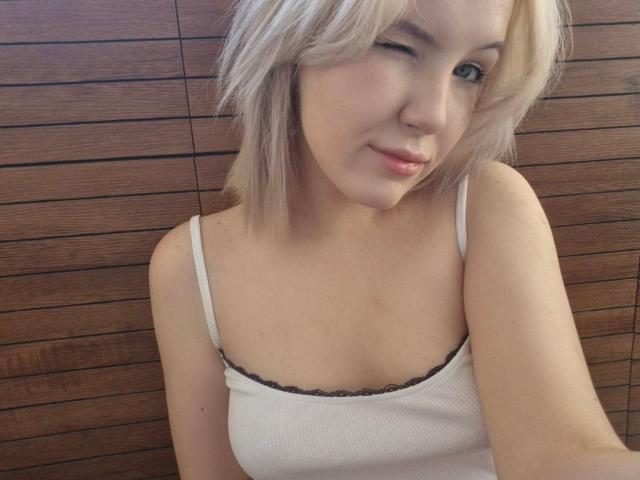 LidiaOckimey with blonde hair, winking, wearing a white tank top showing her small tits with a black lace trim, smiling slightly.