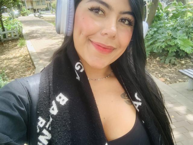 CatalinaReiyna with headphones, black scarf, and red lipstick, smiling at the camera, showing her tits and a small tattoo under her collarbone, in a park.