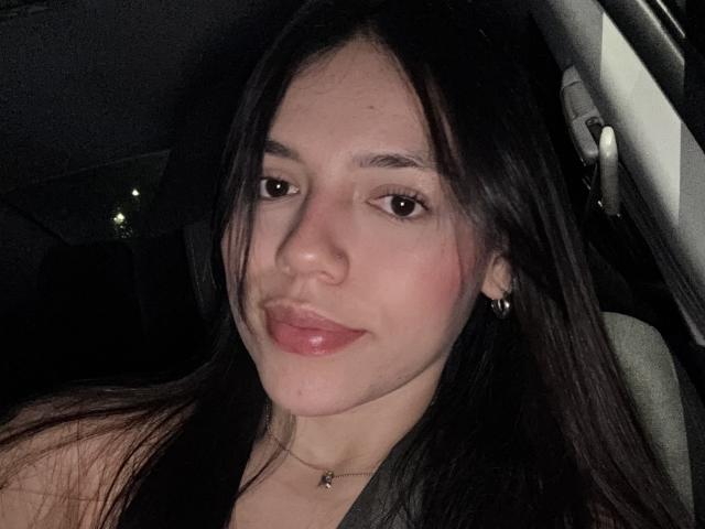 SophiaMarx has shiny pink lips, brown eyes, and long black hair in a car at night, wearing a necklace and small earrings, with a slight smile.