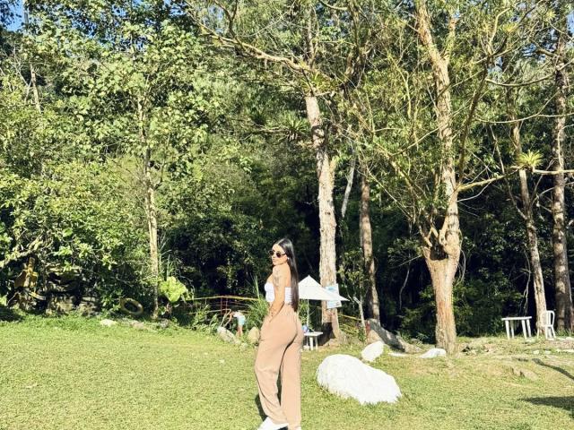 SophiaMarx stands in a sunlit grassy area, wearing tan pants and a white crop top, with her ass facing the camera, looking back over her shoulder.