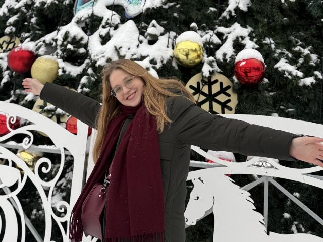 ColettaCorman com os seios à mostra, braços estendidos, sorrindo, usando óculos, casaco verde, cachecol vermelho, neve na árvore, cerca branca ornamentada, enfeites de Natal, segurando uma bola dourada..