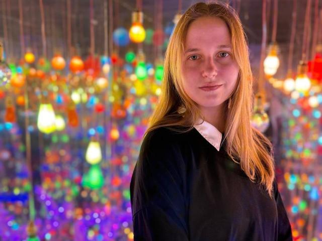 Coletta Corman, com cabelos loiros longos, vestindo uma camisa preta, tem um rosto bonito e seios pequenos, e está em uma sala colorida, iluminada com bokeh, com luzes penduradas..