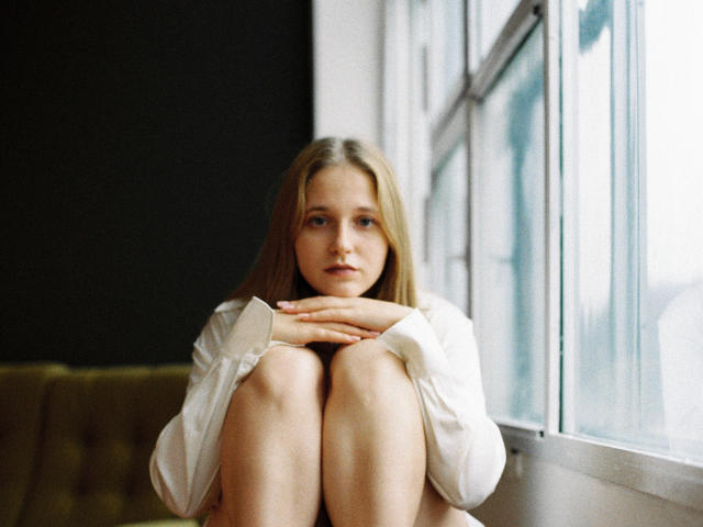 ColettaCorman sitzt mit angezogenen Knien und Händen auf den Knien, trägt ein übergroßes weißes Hemd und blickt mit einem sanften, nachdenklichen Ausdruck aus einem Fenster. Sie hat blondes Haar und helle Haut..