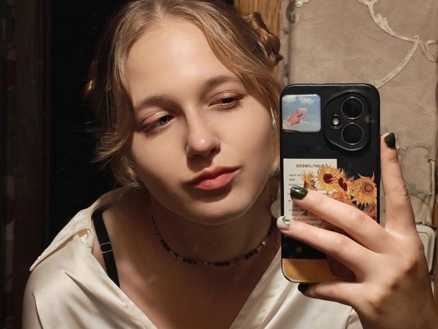 ColettaCorman mit heller Haut, blonden Haaren und grünem Nagellack, macht ein Selfie mit einer Handyhülle mit einem Sonnenblumenmotiv, trägt ein weißes Hemd und einen schwarzen BH, in einem schwach beleuchteten Raum..