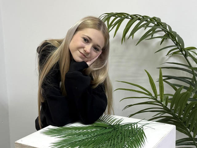 VeraBein, avec de longs cheveux blonds, un sweat à capuche noir, appuyée sur une table blanche, souriante, la tête dans la main, des feuilles de palmier vertes devant et derrière, lumière naturelle douce, détendue, mignonne..