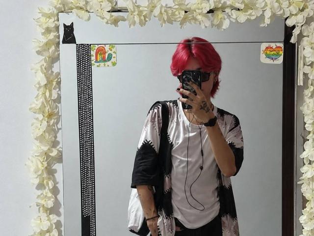 Selfie en el espejo de KaiaFinn con cabello rojo, vistiendo una camisa con estampado blanco y negro, una camiseta blanca y pantalones negros, sosteniendo un teléfono que cubre su rostro, marco de flores blancas..