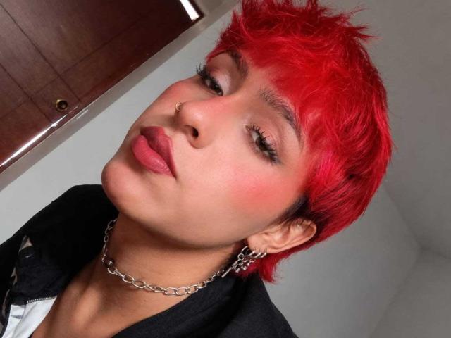 KaiaFinn, com cabelo curto, ruivo e bagunçado, piercing no nariz e colar, vestindo uma jaqueta escura, batom rosa e brincos de corrente, olhando para cima com um leve sorriso..