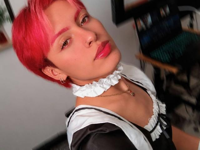 KaiaFinn, com cabelo rosa curto, piercing no nariz, batom rosa, uniforme de empregada preto e branco, colar com babados, colar de ouro e um sorriso confiante. Seus seios estão ligeiramente visíveis..