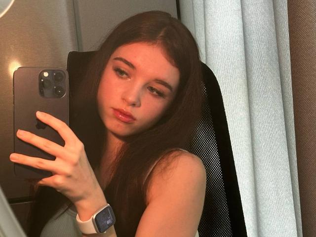 ChuTristan with long brown hair, pouty lips, and a serious expression, holding a phone, wearing a white wristband, sitting in a black chair, taking a selfie.