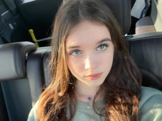 ChuTristan, con cabello castaño largo, ojos azules y un pequeño collar azul, sentada en un coche, mirando directamente a la cámara, vestida con una camisa verde, con un rostro pálido..