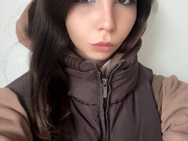 VickiLipoma hat dunkelbraunes Haar, hellbraune Haut und volle, rosafarbene Lippen. Sie trägt eine braune, gesteppte Weste über ein beiges Hemd. Ihre Augen sind zur Seite gerichtet..