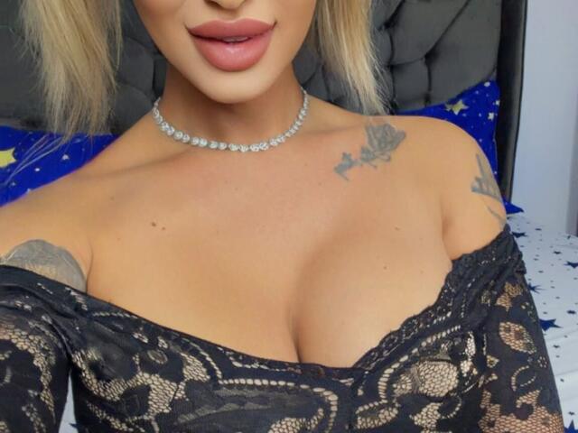 NicolSNaya, con cabello rubio, vistiendo una blusa negra de encaje con un hombro descubierto, mostrando sus senos, un collar de perlas y tatuajes en el pecho y el hombro, sonriendo con labios rosados..