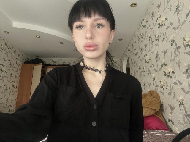 DawnPrinty with pale skin, black bangs, and green eyes, wearing a black shirt and black choker, pouting pink lips, showing small tits, in a floral-patterned room.