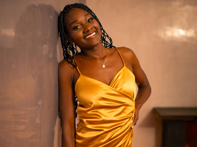 AndreaStorm, smiling, wearing a shiny yellow satin dress, hands on hips, braided hair, dark skin, gold necklace, standing against a wall, looking happy.