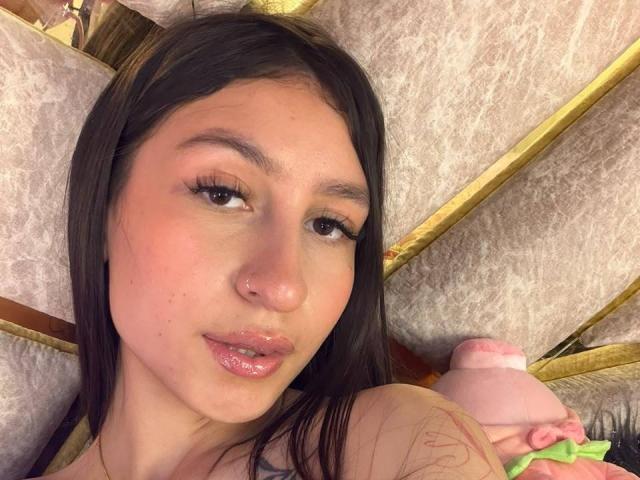 MaddyCarina, con le labbra rosa lucide, tiene in mano un dildo rosa, ha i capelli castano scuro, un piccolo piercing al naso e un leggero rossore sulle guance. I suoi occhi sono leggermente aperti..