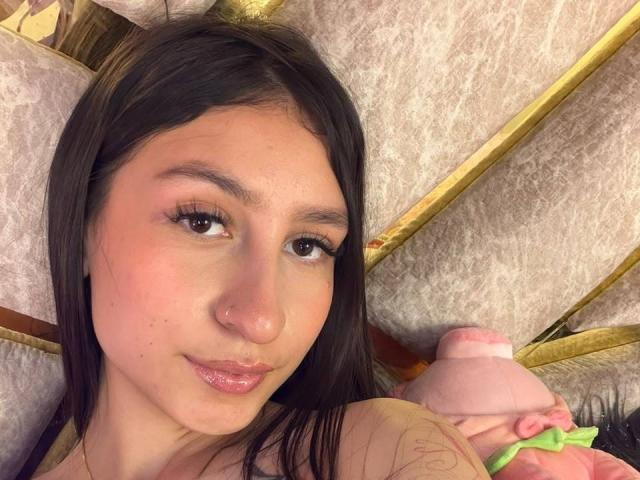 MaddyCarina, avec des lèvres roses, des cheveux noirs et des yeux marrons, regardant l'appareil photo, allongée sur un lit avec un animal en peluche rose et des oreillers dorés..