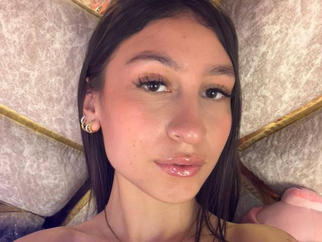 MaddyCarinas Gesicht, mit glänzenden rosafarbenen Lippen und einer kleinen Nase, hat glattes, braunes Haar, braune Augen und trägt ein goldenes Ohrring. Sie liegt auf einem strukturierten Kissen..