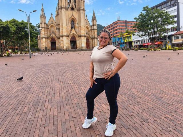 Vianca Castellanos è in piedi con sicurezza, con le mani sui fianchi, indossa una maglietta beige, pantaloni neri e scarpe da ginnastica bianche, di fronte a una cattedrale gotica, circondata da piccioni su una piazza in mattoni..