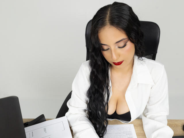 StefanyDelly, con cabello negro largo, lápiz labial rojo, bata de laboratorio blanca, sostén negro, senos grandes, mirando hacia abajo un portátil y documentos sobre un escritorio de madera, sentada en una silla de oficina negra..