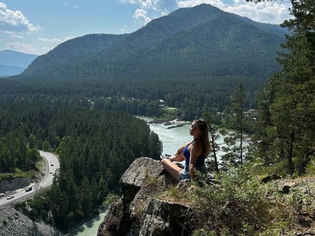 BernardineShader está sentada en un saliente rocoso, vestida con una camiseta azul y pantalones cortos, con el trasero mirando hacia el río, mirando hacia atrás la carretera sinuosa y las montañas boscosas..