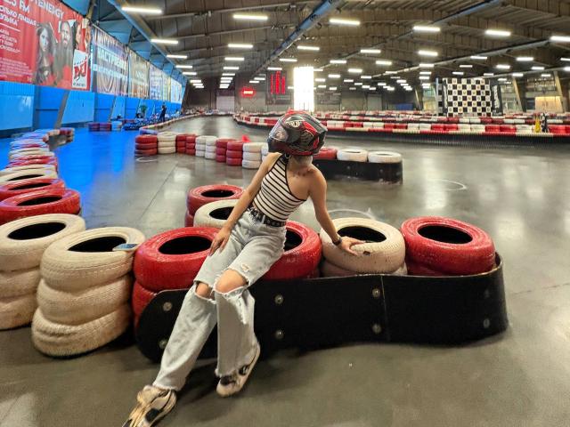 BernardineShader, die ein gestreiftes Oberteil und zerrissene Jeans trägt, sitzt auf roten Reifen in einer Garage, mit einem gemusterten Helm und Rollschuhen, und blickt leicht zur Seite..