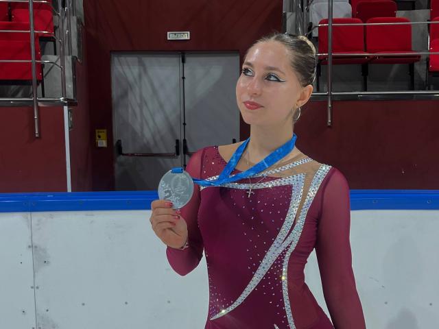 BernardineShader tiene una medalla de plata, lleva un traje de gimnasia brillante color burdeos y plateado, con un lazo azul, en una pista de hielo, sonriendo, con el cabello castaño recogido en un moño apretado..