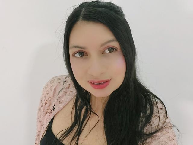 Ghaby, avec de longs cheveux noirs, des yeux roses et des lèvres rouges, portant un cardigan en dentelle rose par-dessus des seins nus, regardant l'appareil photo avec un léger sourire..