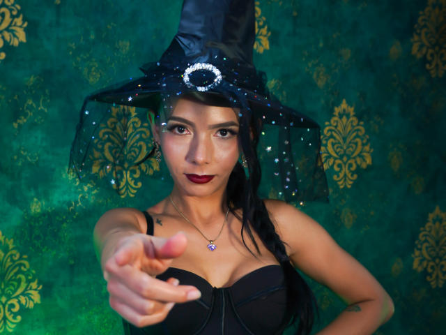 EvelinLuna, avec un chapeau de sorcière noir, pointant vers la caméra, portant un soutien-gorge noir, du rouge à lèvres rouge, une longue tresse noire, un petit collier rose, un fond vert avec des motifs dorés. Seins, mignonne, confiante..