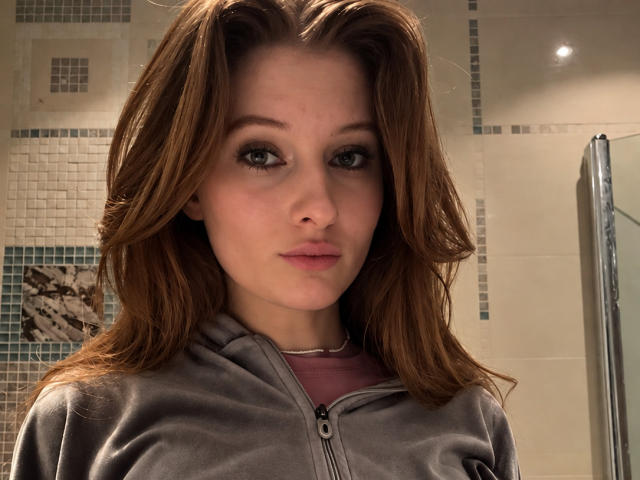 ArdelleCooksey ha i capelli castani, gli occhi verdi e le labbra rosa, indossa una felpa grigia sopra una maglietta rosa, con un'espressione neutra, in un bagno..