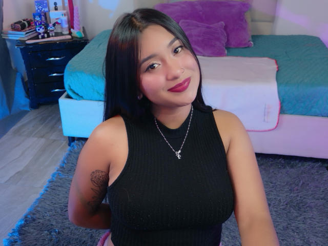 HannahJohnes, met donker haar, draagt een zwart, geribbeld tanktopje, waarbij haar borsten zichtbaar zijn. Ze heeft een tatoeage op haar linkerarm en draagt een ketting. Ze zit voor een bed met turquoise en roze kussens..