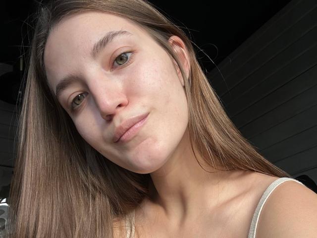 MayPayer has light green eyes, straight brown hair, and is smiling slightly with her pouty lips, showing her smooth, fair-skinned face and small tits in a white strap.