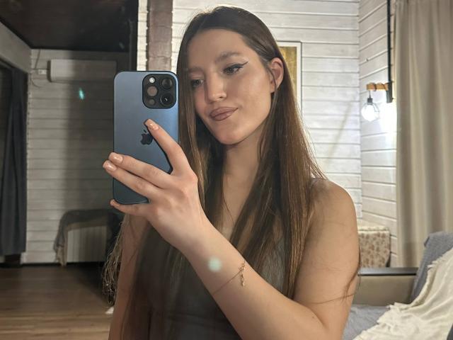 MayPayer with long brown hair, holding a blue iPhone, taking a selfie in a mirror. She has small tits, subtle makeup, and a slight smile. Wood floor, white walls.
