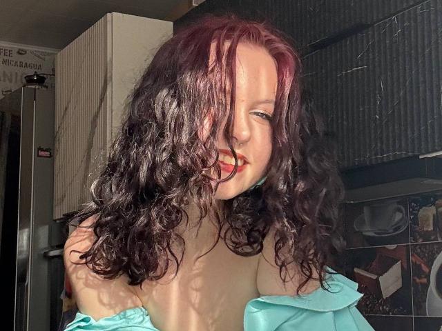 Sherita Pinales, met krullend zwart haar, rode highlights en een schattige glimlach, draagt een lichtblauwe top met een open schouder, waardoor haar borsten zichtbaar zijn, in een keuken met veel zonlicht..