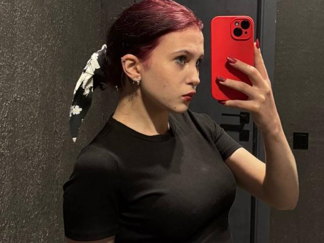 SheritaPinales met rood haar in een staart, een zwart T-shirt, die een rode telefoon vasthoudt, een spiegel-selfie maakt, kleine borsten laat zien, bleke huid, rode nagellak, en een serieuze blik heeft..