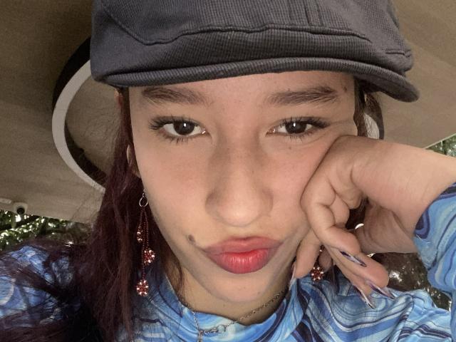 JuliethCourtouis with a grey flat cap, pouty red lips, dark eyebrows, and a small mole on her left cheek. She's wearing a blue plaid shirt and pink earrings.