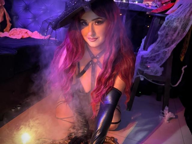MiaRouusee met lang rood haar, een zwarte leren hoed en zwarte, open lingerie, draagt lange zwarte handschoenen, knielt op een tafel met rook en glimlacht verleidelijk..