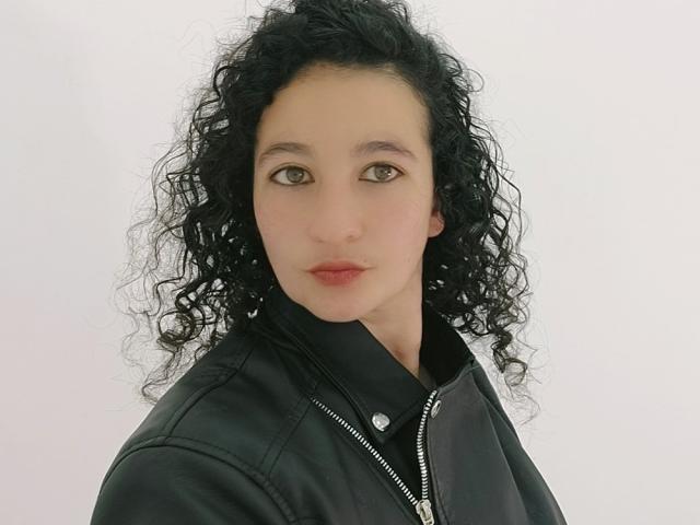 ShamantaJ hat lockiges, schwarzes Haar, helle Haut, braune Augen und trägt eine schwarze Lederjacke mit einem Reißverschluss. Sie schaut direkt in die Kamera und hat einen neutralen Gesichtsausdruck..