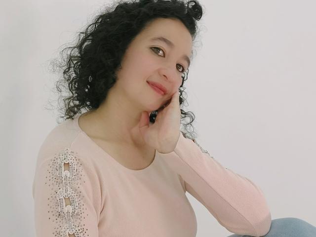 ShamantaJ, con cabello negro rizado, piel pálida y un suéter rosa con mangas ornamentadas, apoyando su barbilla en la mano, sonriendo suavemente. Sus pechos son pequeños y es linda..