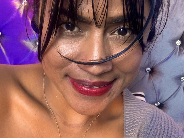 IvannaDuran69 has dark hair, red lipstick, and a slight smile, with a single black hair strand across her nose. She's wearing a gold necklace, and has a tan skin tone.