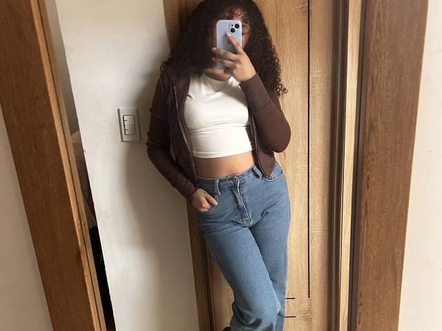 SofiaPassionne, con i capelli ricci, indossa una giacca marrone, un top corto bianco e jeans blu, e sta scattando un selfie allo specchio, mostrando i suoi glutei sodi e una mano in tasca..