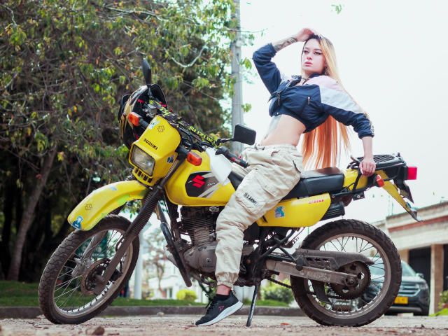 MiaValenttine with long blonde hair, wearing a blue jacket, tan cargo pants, and black sneakers, sitting on a yellow motorcycle, hand in hair, looking at camera.