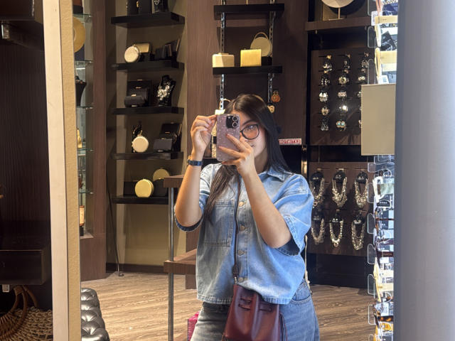 MiiaPalmer with long black hair, wearing glasses and a denim shirt, taking a mirror selfie in a jewelry store, holding a phone with a colorful case, brown purse.