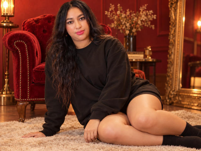 Una ragazza latina con lunghi capelli neri, che indossa un maglione nero e calze nere, seduta su un tappeto con le gambe incrociate, che mostra il suo sedere scoperto, in una stanza accogliente di colore rosso..