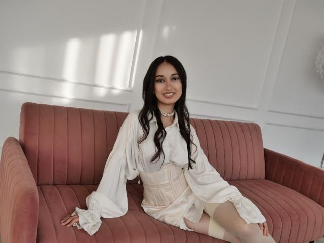 ElviraWesdety sits on a pink ribbed couch, wearing a white long-sleeve dress and corset, smiling with long black hair, showing her white thigh-high socks.